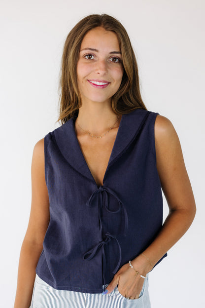 Young woman wearing a navy sleeveless top that is loose fitting and has a 2 tie front closure.