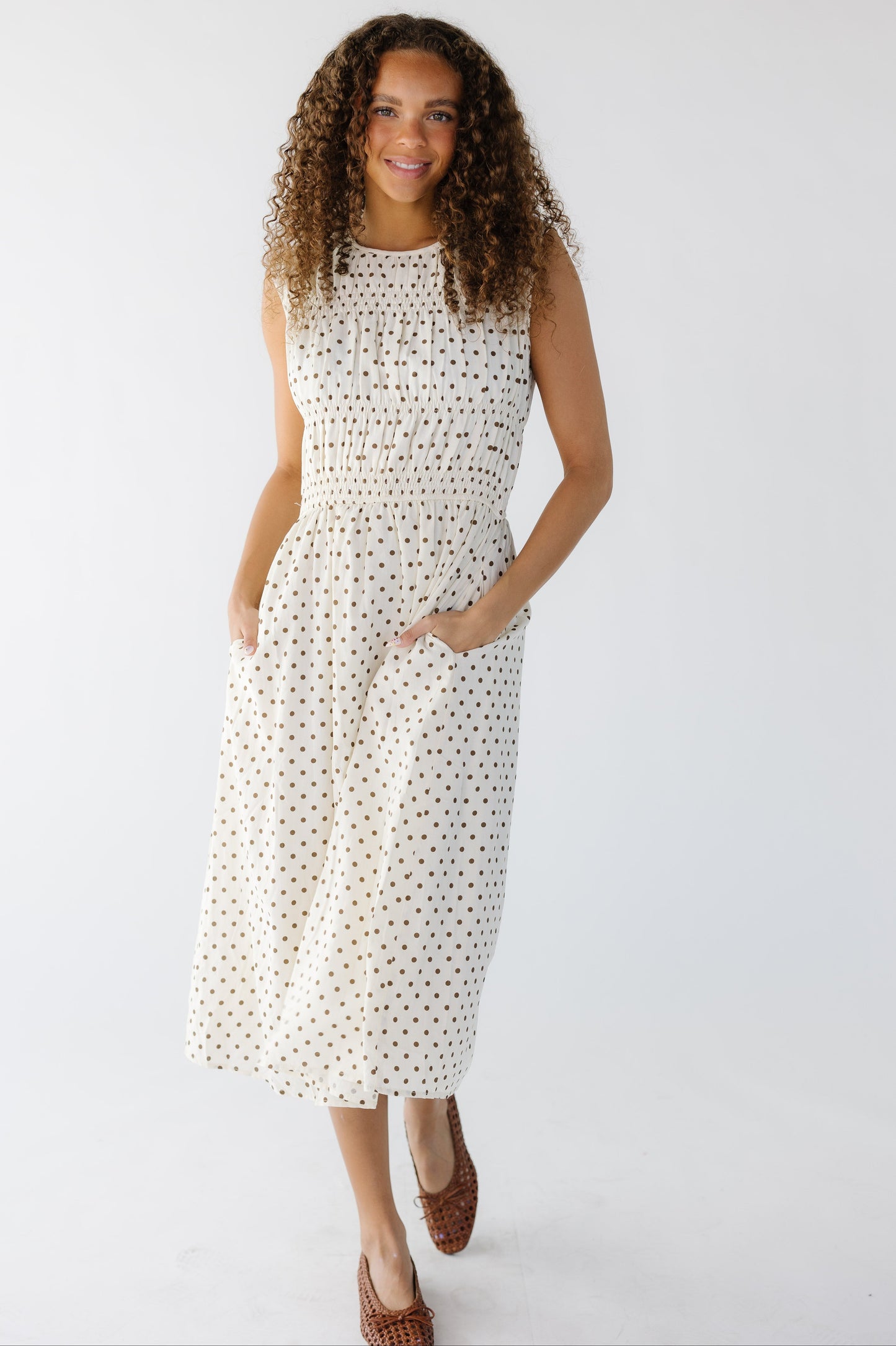 Teenage girl wearing a  sleeveless cream and brown polka dot midi dress with side pockets and groupings of smocked rows on the bodice with a round neckline.