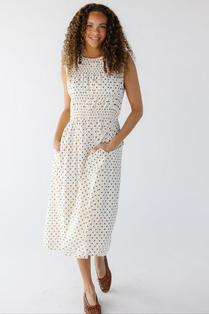 Teenage girl wearing a  sleeveless cream and brown polka dot midi dress with side pockets and groupings of smocked rows on the bodice with a round neckline.