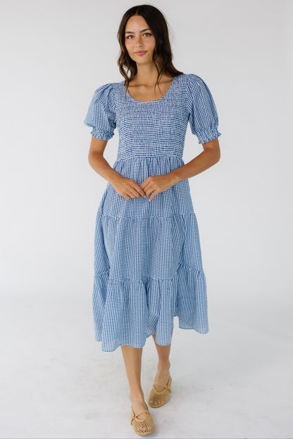 Young woman wearing a blue gingham midi dress with short puff sleeves, a scoop neckline and multiple gathered tieres.