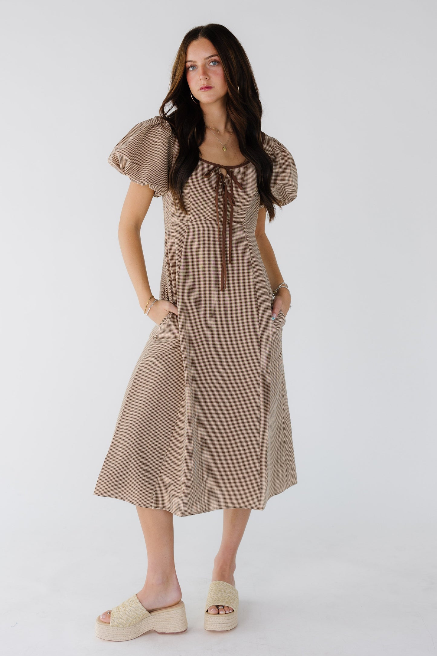 Young woman wearing a brown gingham midi dress with balloon sleeves a close fitting bodice, and a wide scoop neckline with 2 front ties ata the neckline.