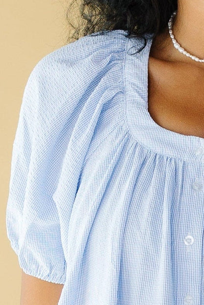 Close view of a woman wearing a light blue line plaid button down dress with a scoop neckline and balloon sleeves.
