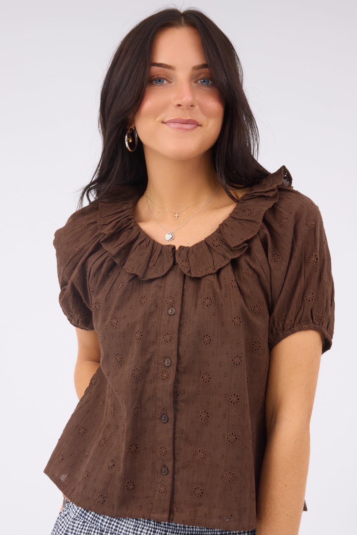 Woman wearing a loose fitting brown eyelet blouse that buttons down the front with a ruffled neckline.