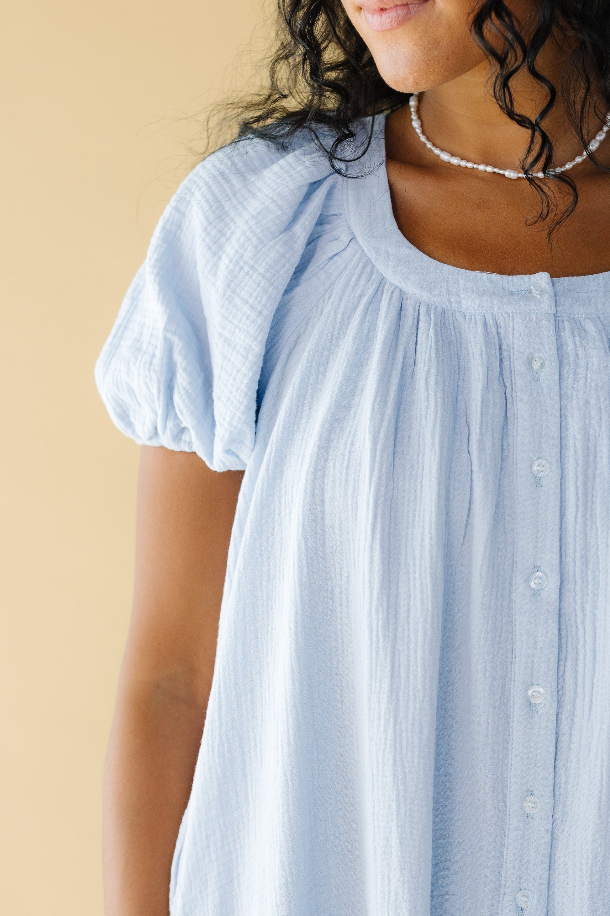 Close view of a woman wearing a light blue gauzy button down dress with large puffy sleeves and a scoop neckline.