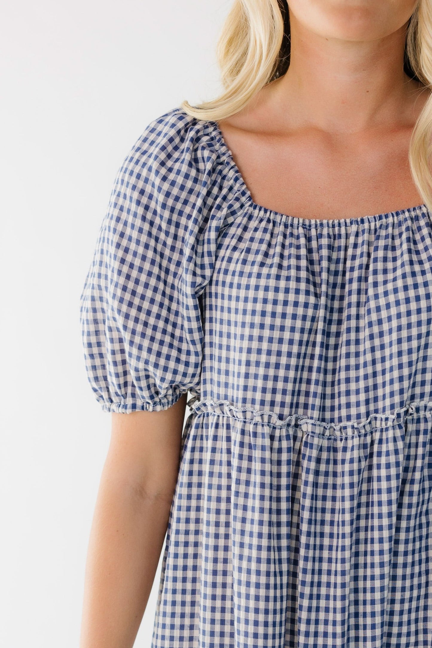 Close view of a woman wearing a blue & white gingham tiered dress with large puff sleeves and an elastic square neckline.