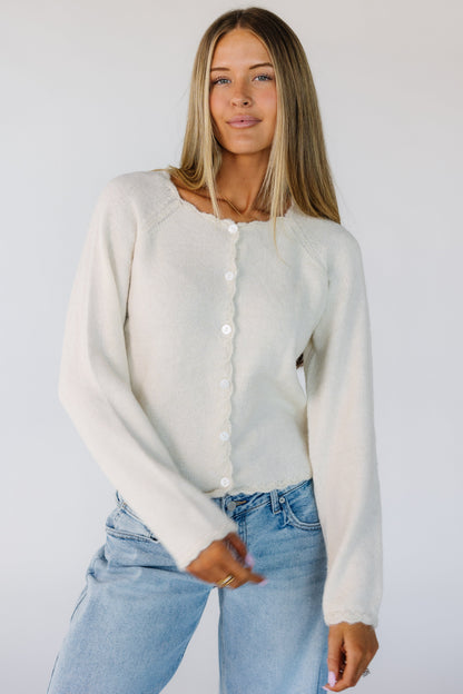 Woman wearing a modest cream cardigan with scallop edge, round neckline and long sleeves with scallop cuffs.