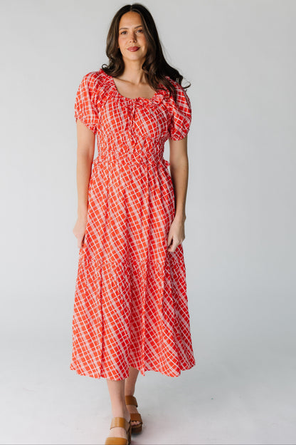 Woman wearing a modest red plaid midi dress with short puff sleeves, a wide close fitting waist and a wide bottom ruffle.