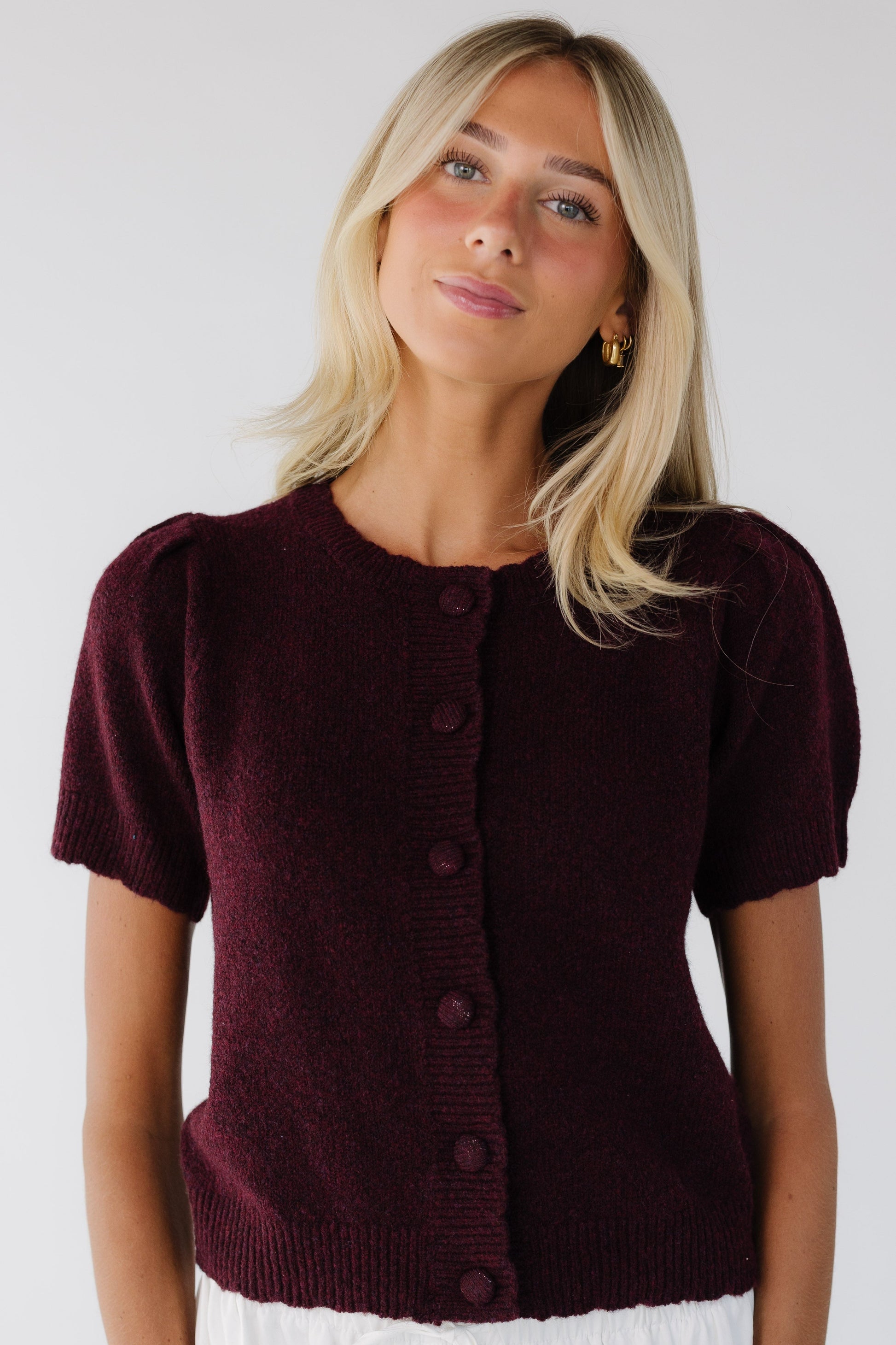 Woman wearing a burgundy button down cardigan with matching covered buttons, scallop hems and crew neckline, and short sleeves.