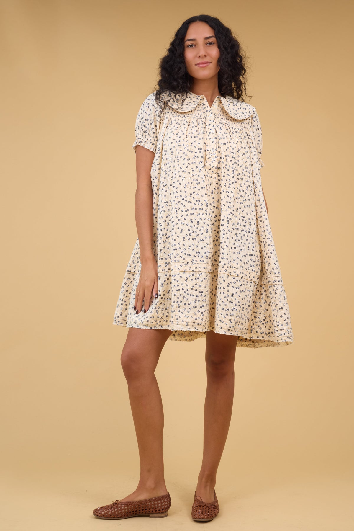 Woman wearing a short cream dress with blue floral print, short sleeves, short button placket, smockedyoke and a wide round collar.