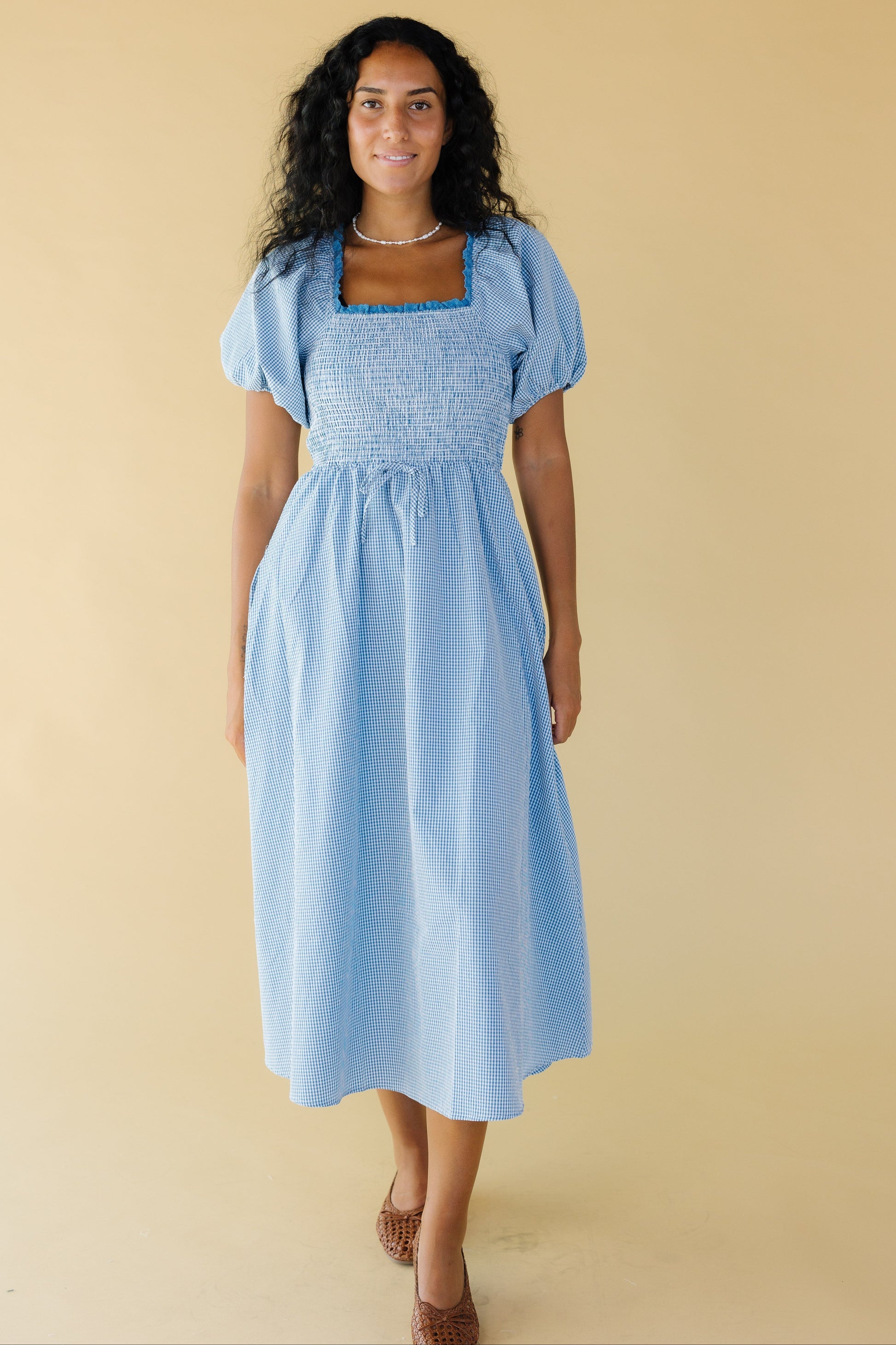 Woman wearing  blue & white gingham midi dress with with a snug smocked bodice, a lace trimmed square neckline, balloon sleeves and a full skirt.
