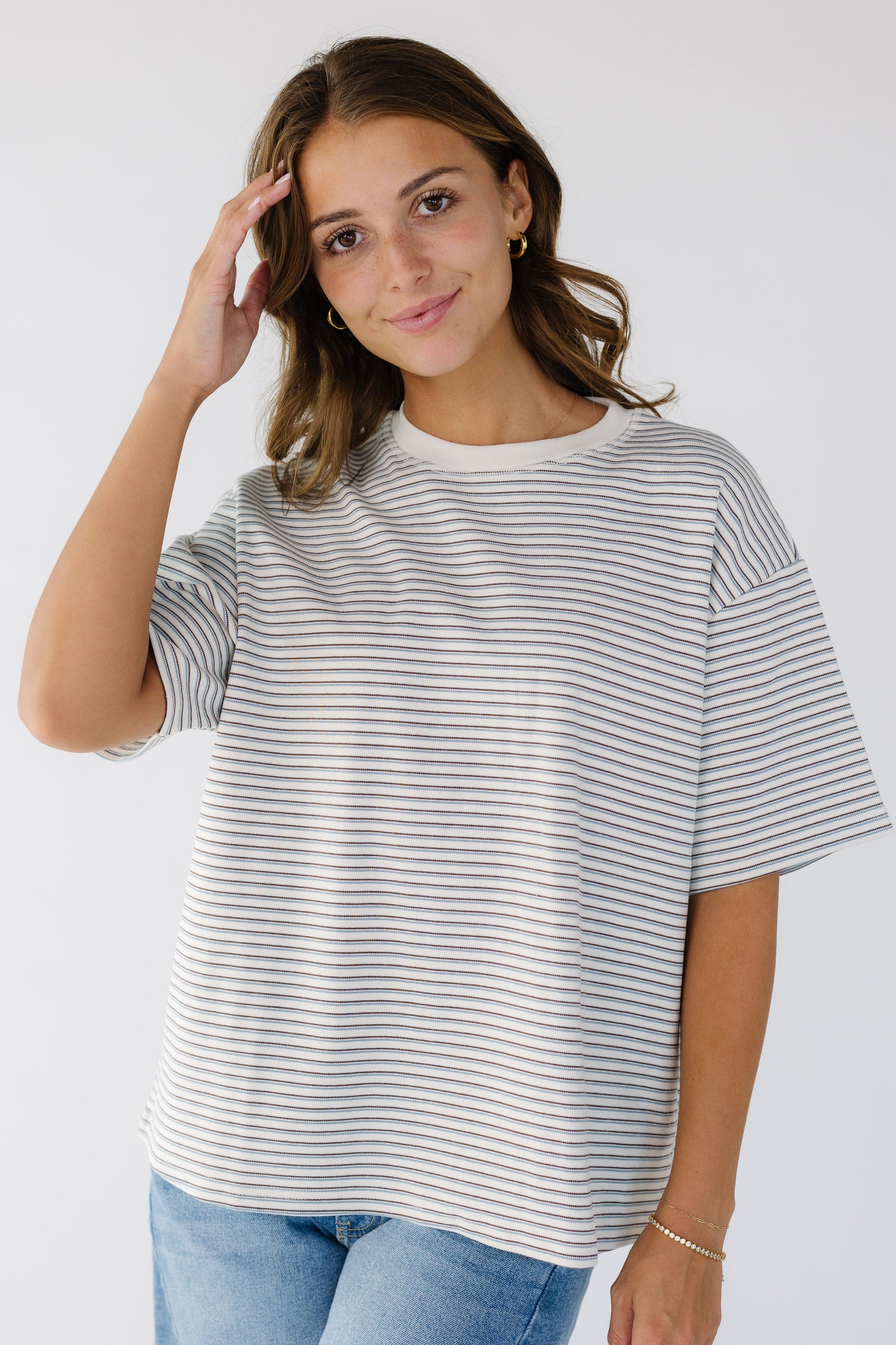 Woman wearing a white loose boxy tee with thin blue & brown stripes with short sleeves ad a white crew neckline.