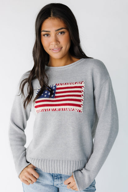 Woman wearing a light grey long sleeve sweater with a large front American flag graphic, long sleeves and rolled hems.