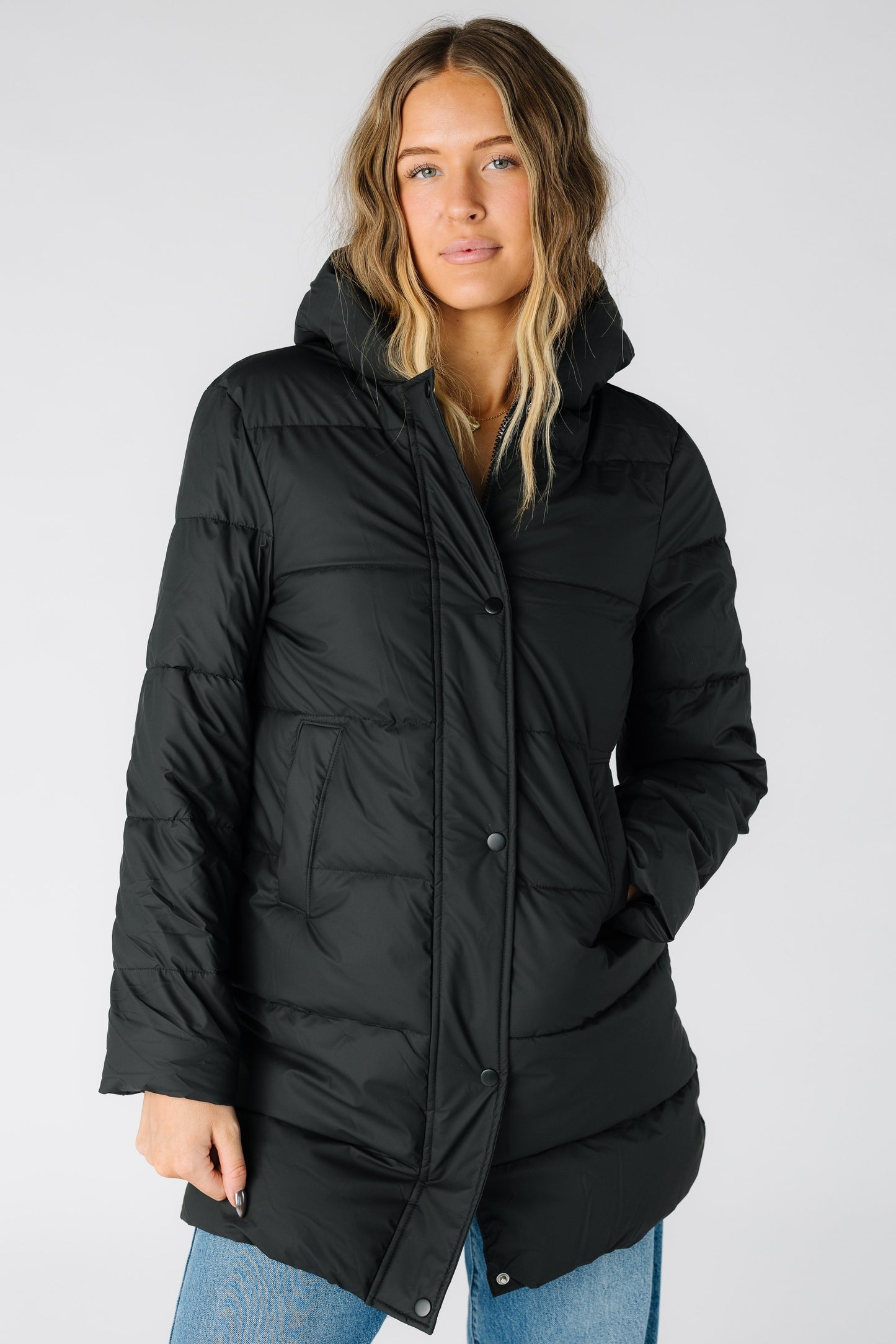 Woman wearing a a long sleeve black puffer jacket with pockets and a puffy hood