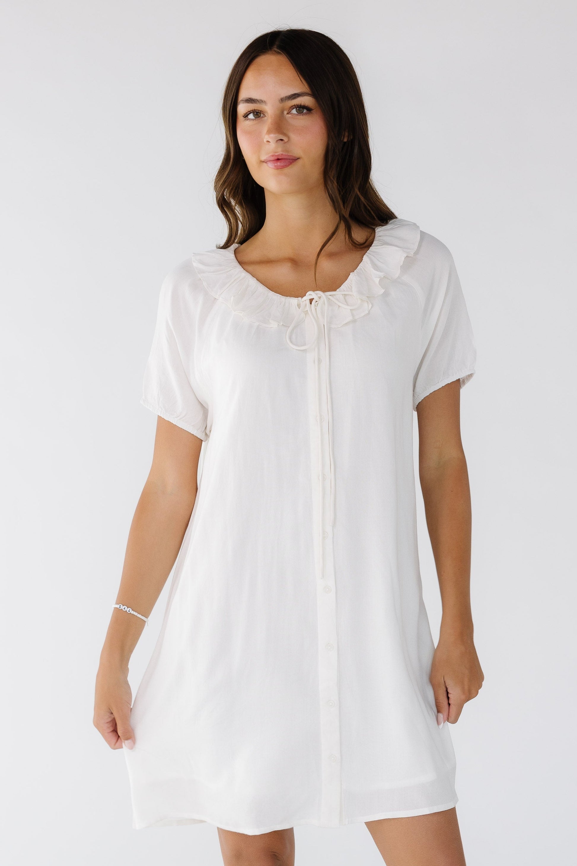 Woman wearing a short white button down dress with short sleeves and a round ruffled neckline with a long front tie.