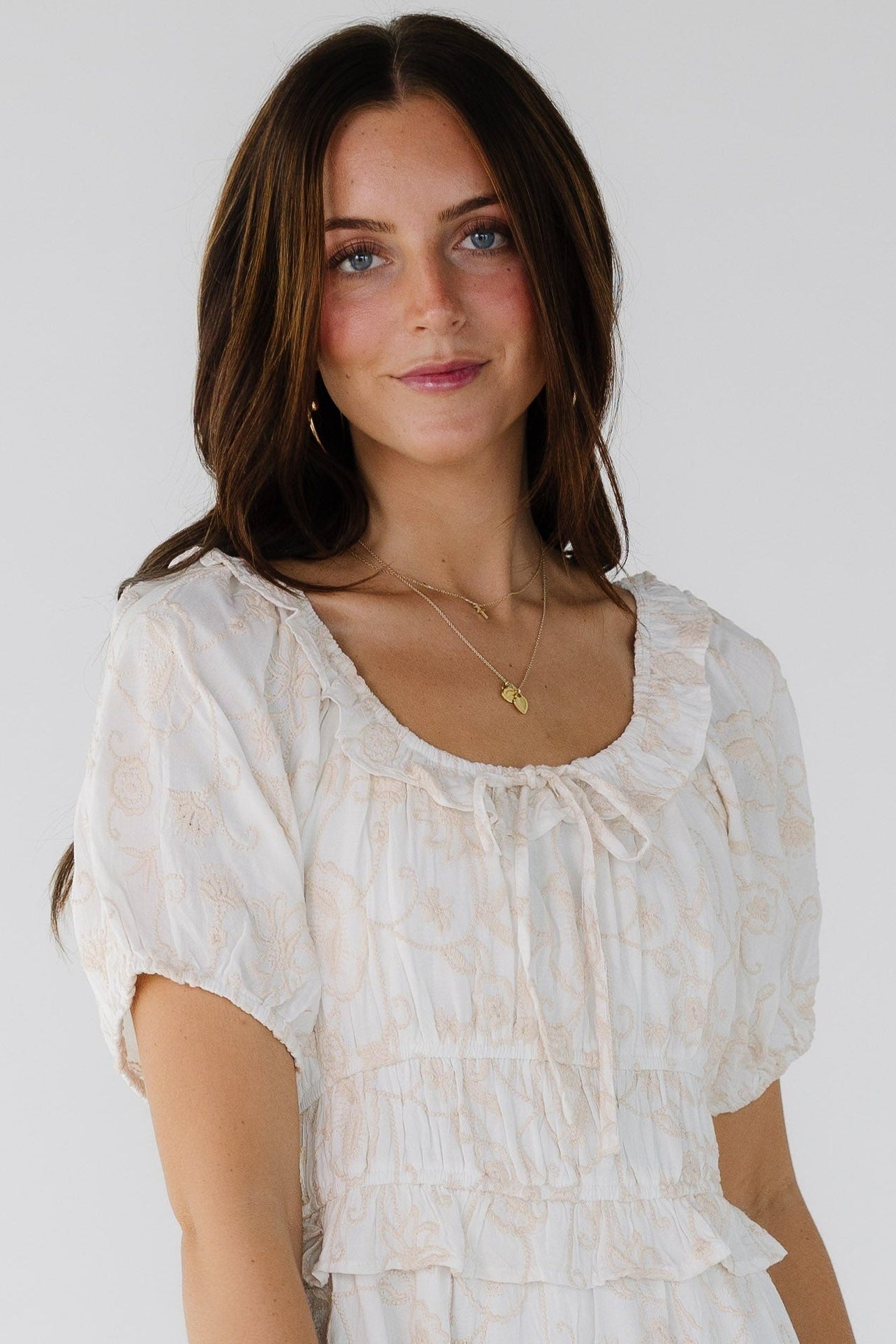 Close view of a woman wearing a white and champagne color floral embroidery with a ruffled scoop neckline, puff sleeves and a wide waist.