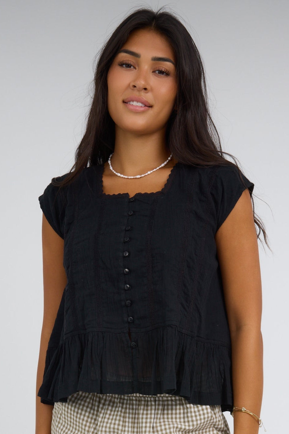 Young woman wearing a black loose fitting button down blouse with a square neckline, cap sleeves and a bottom ruffle.
