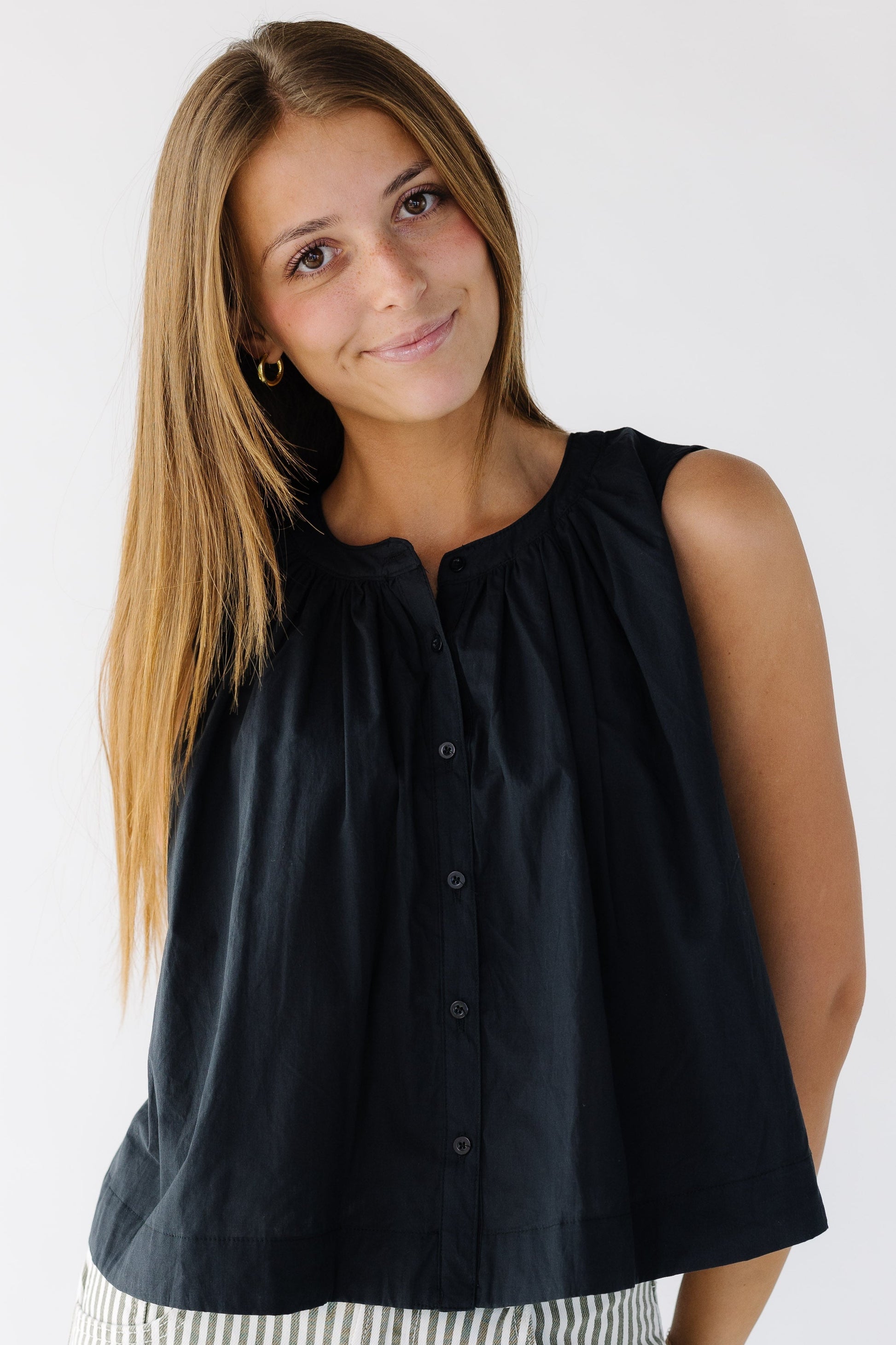 Young woman wearing a sleeveless button down blouse in black that is  loose fitting with a banded round neckline with gathers for the blouse.