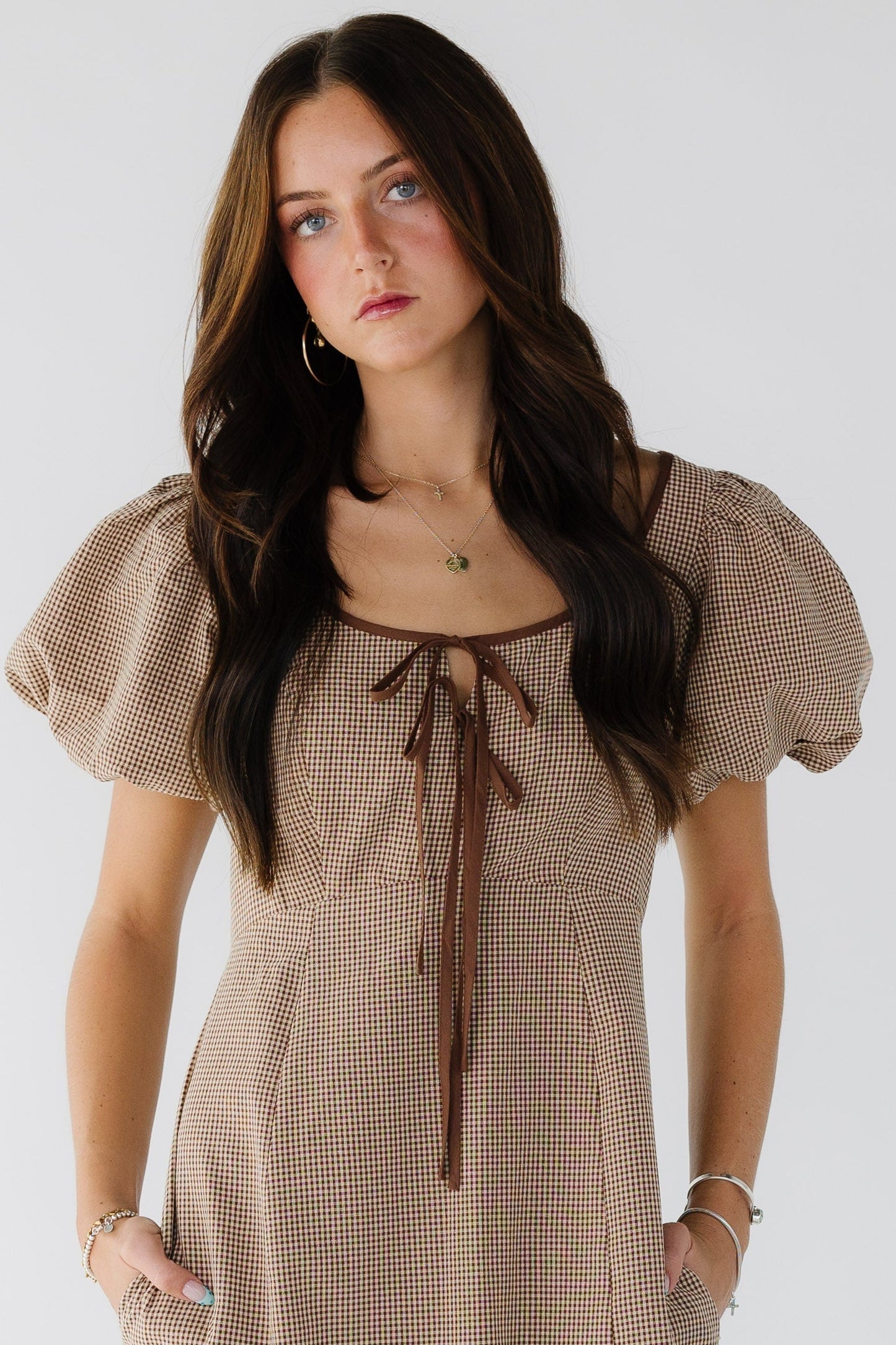 Woman wearing a brown gingham dresswith large balloon sleeves, wide scoop neckline with front bodice ties and side pockets.