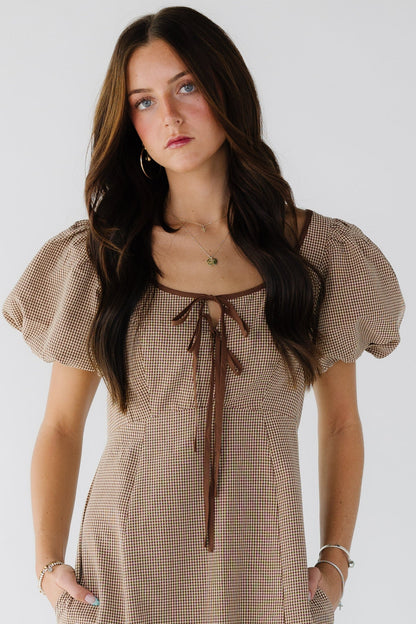 Woman wearing a brown gingham dresswith large balloon sleeves, wide scoop neckline with front bodice ties and side pockets.