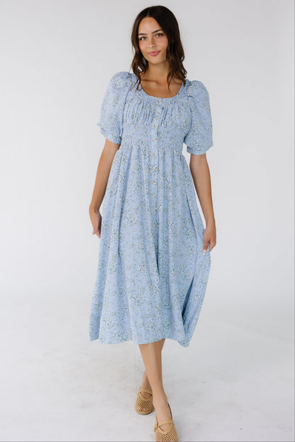 Woman wearing a blue floral button down dress with a scoop neckline, large puff sleeves and a full skirt.