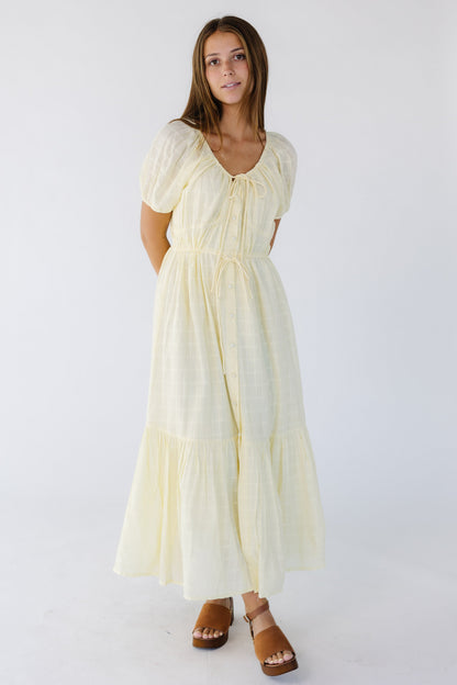 Woman wearing a light yellow button down midi-maxi dress with a wide bottom ruffle, drawstring waist and short puff sleeves.
