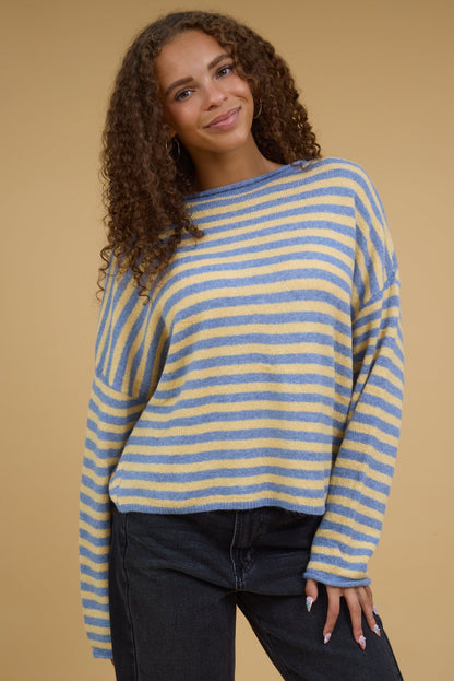 Young woman wearing a loose fitting stripe sweater in yellow & blue stripe with drop shoulders and long sleeves.