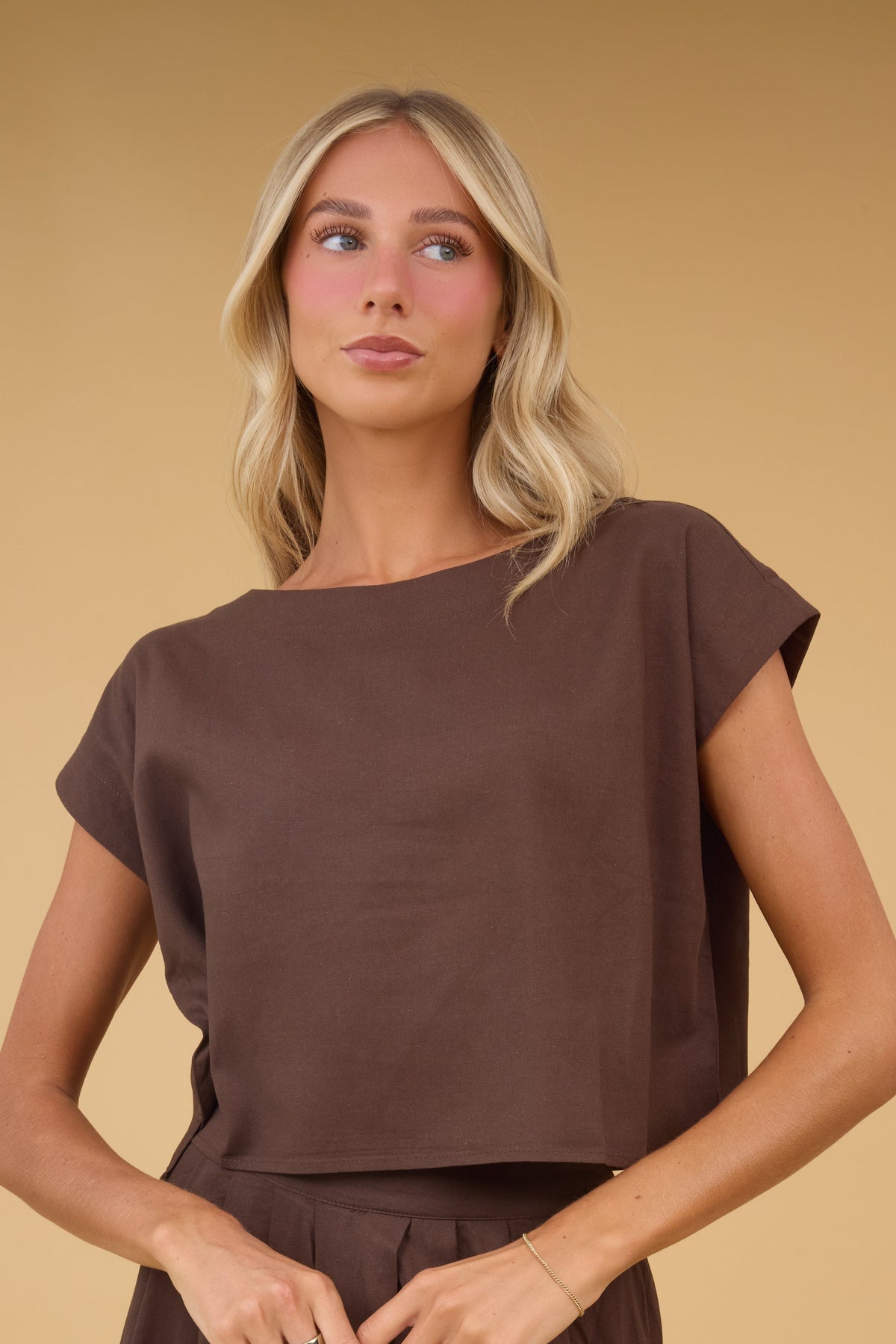 Woman wearing a brown loose fitting blouse with a boat neckline and cap sleeves.