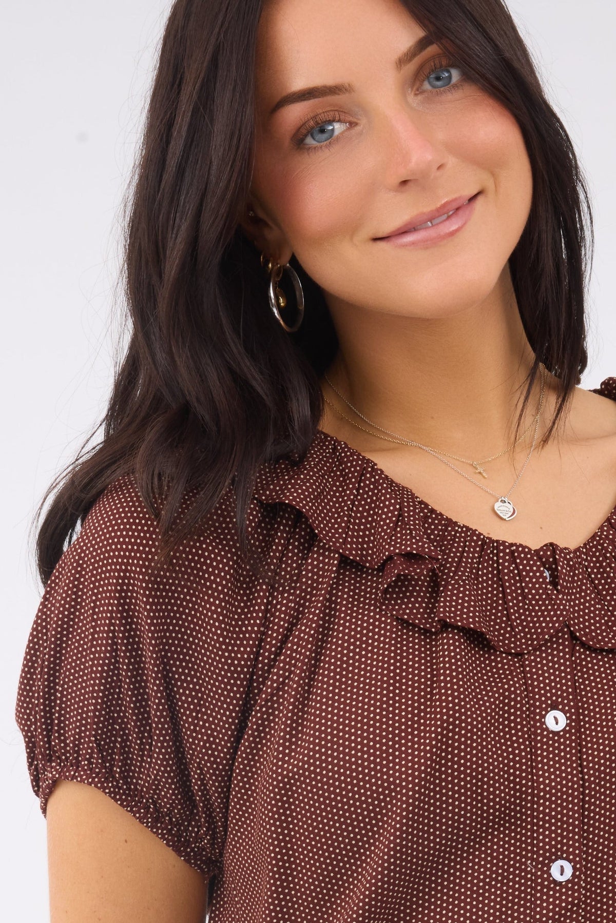A close view of a woman wearing a brown polka dot, button down blouse ith a ruffled collar and short sleeves.