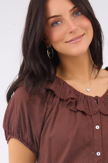 A close view of a woman wearing a brown polka dot, button down blouse ith a ruffled collar and short sleeves.