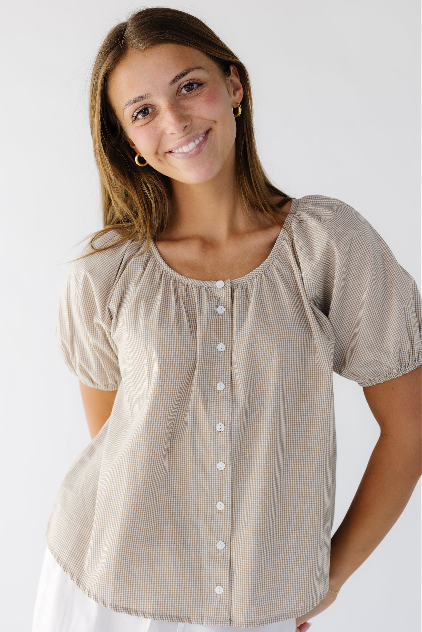 Woman wearing a light brown gingham blouse with a scoop neckline and short sleeves that buttons down the front. 