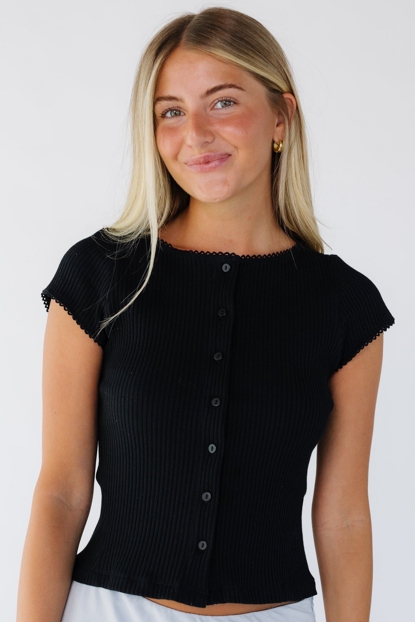 Young woman wearing a modest black button down top that is rib knit and a round neckline and cap sleeves.