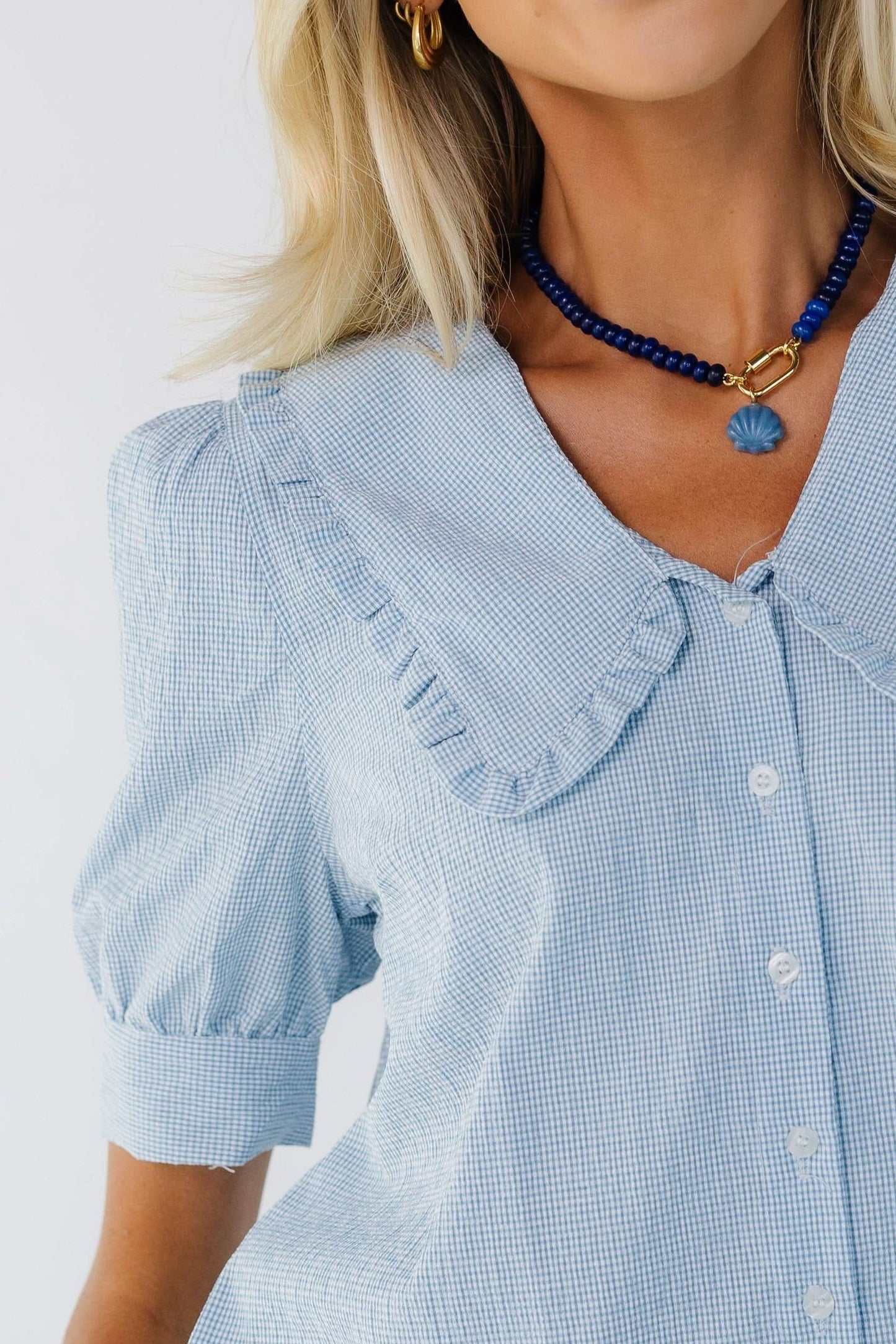 Close view of a woman wearing a textured light blue gingham button down blouse with puff sleeves and a wide, ruffle edge collar on a V-neckline.