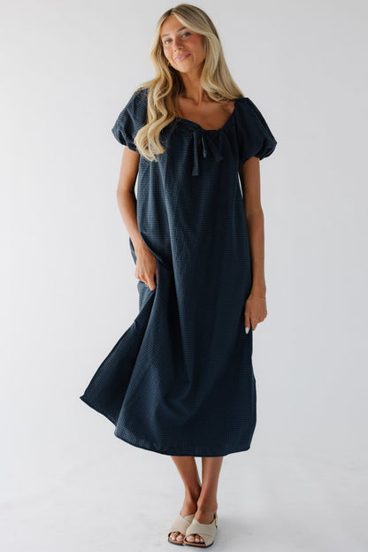 Young woman wearing a modest navy gingham midid dress with large puff sleeves and a drawstring-scoop neckline.
