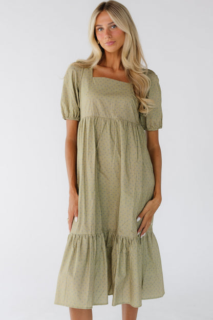 Young woman wearing an olive midi dress with a wide bottom ruffle, short puff sleeves, square neckline and a gathered waistline.