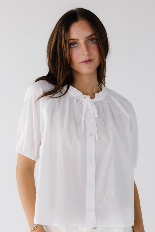 Young woman wearing a modest white button down blouse with white puff sleeves and a ruffled collar and tie.
