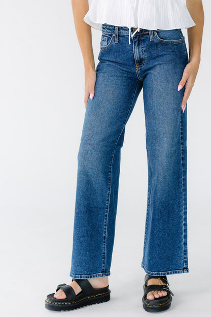 Young woman wearing dark denim jeans with a mid rise waist, wide cut legs and sewn hems.