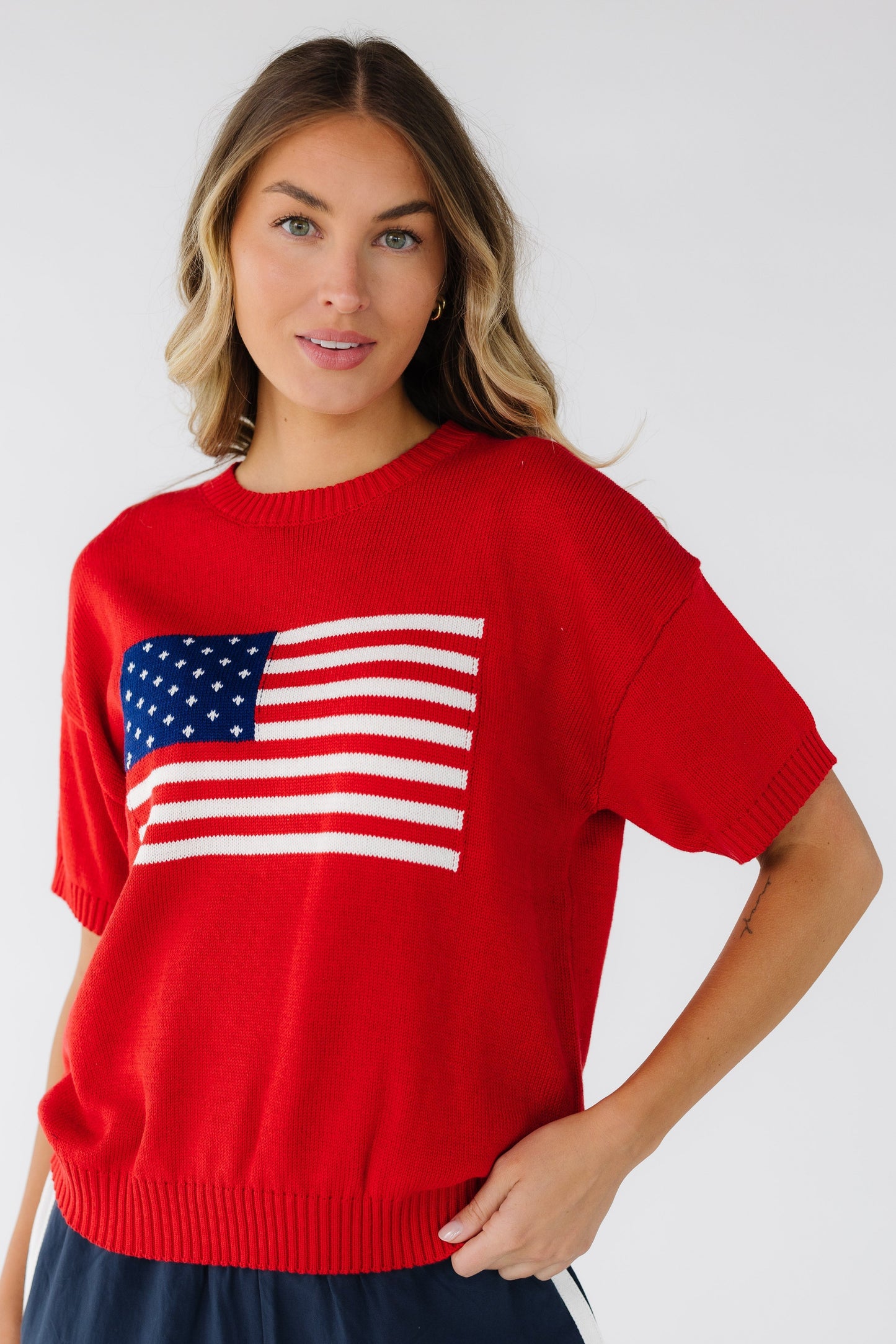 Woman wearing a red short sleeve sweater with a crew neckline, ribbed waist and large front American flag graphic.