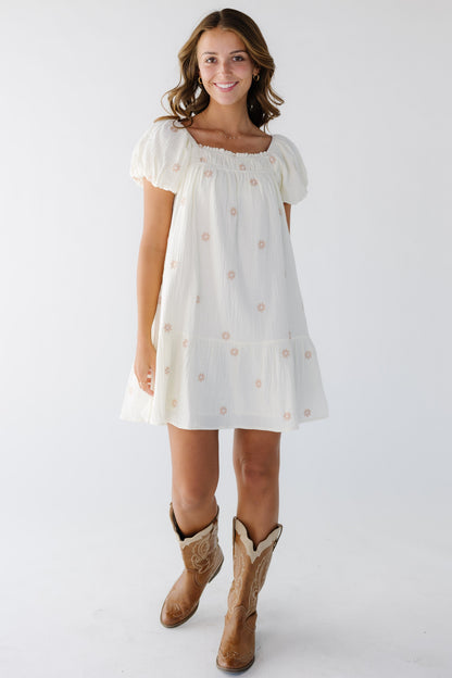 Woman wearing a modest ivory short dress with scattered pink floral embroidered flowers, a square neckline and short puff sleeves.