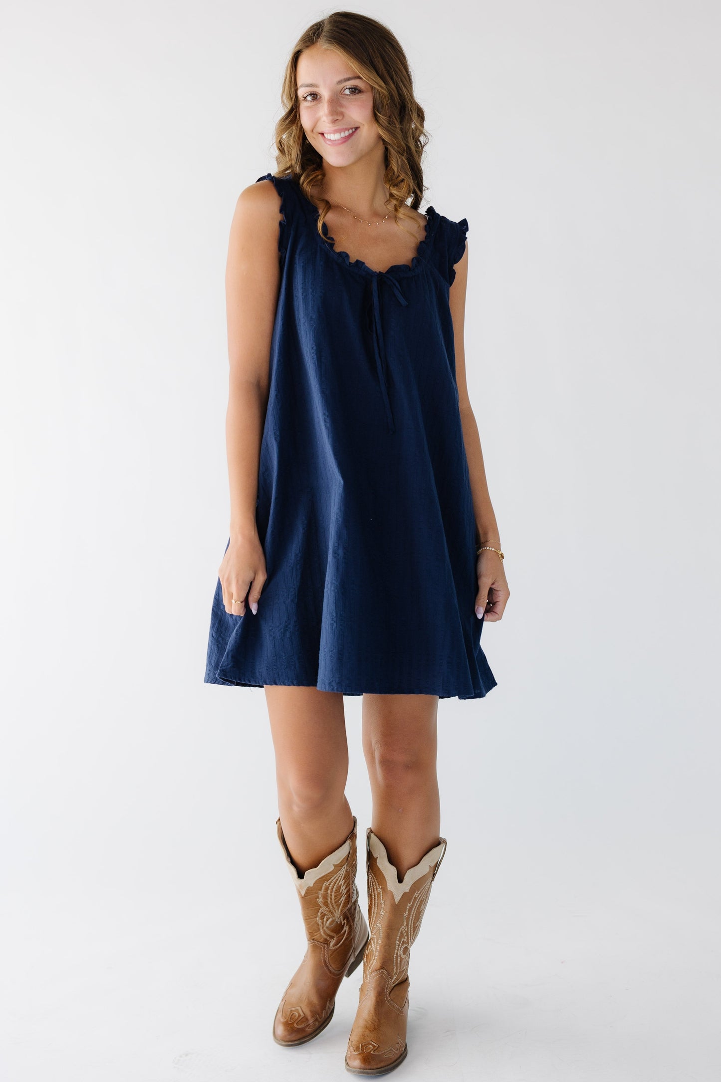 Young woman wearing a navy above the knee swing dress with a ruffled neckline and armholes.