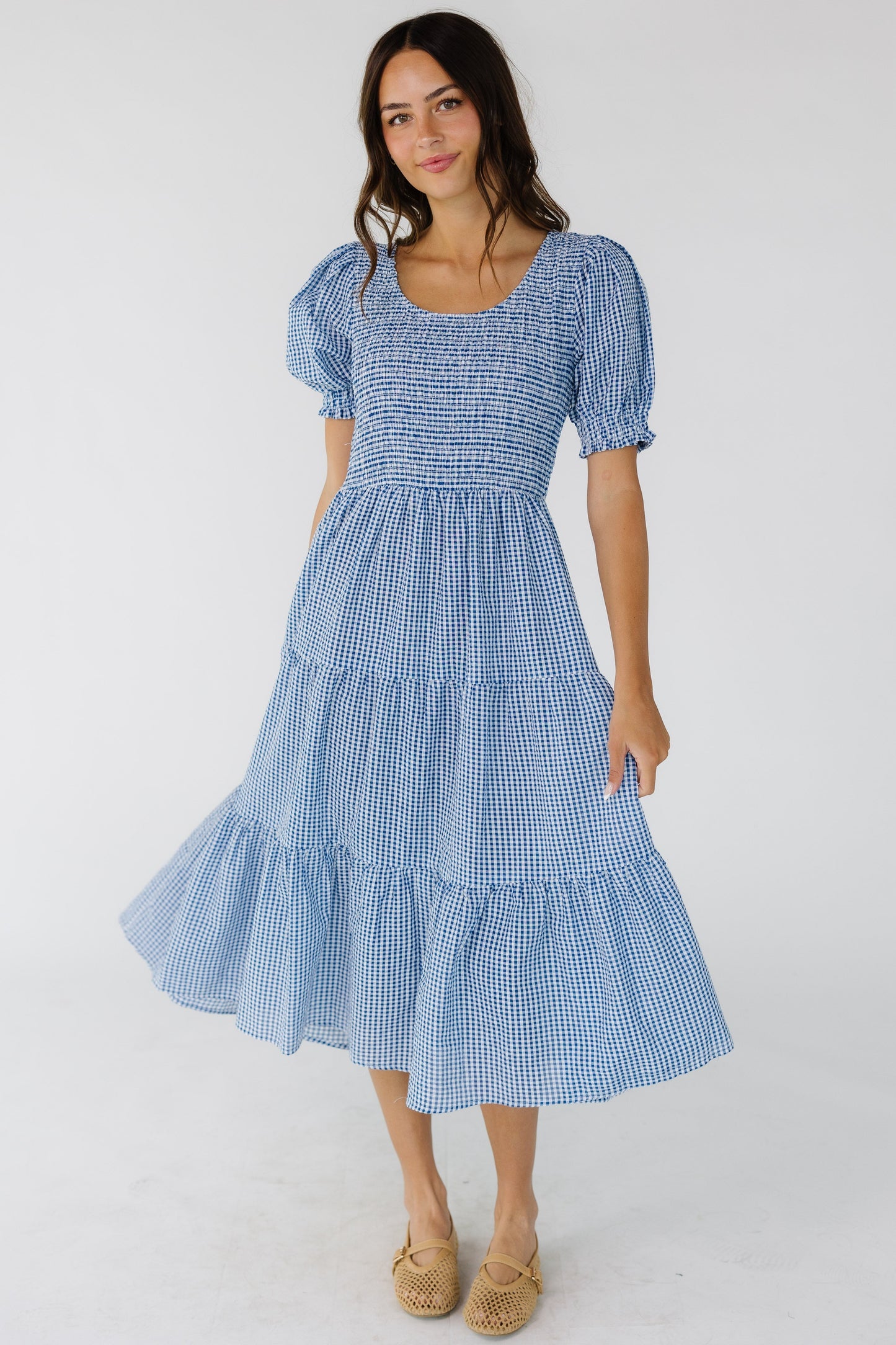 Woman wearing a blue & white gingham midi dress with multiple gathered tiers, smocked front & back bodice, scoop neckline and short puff sleeves.