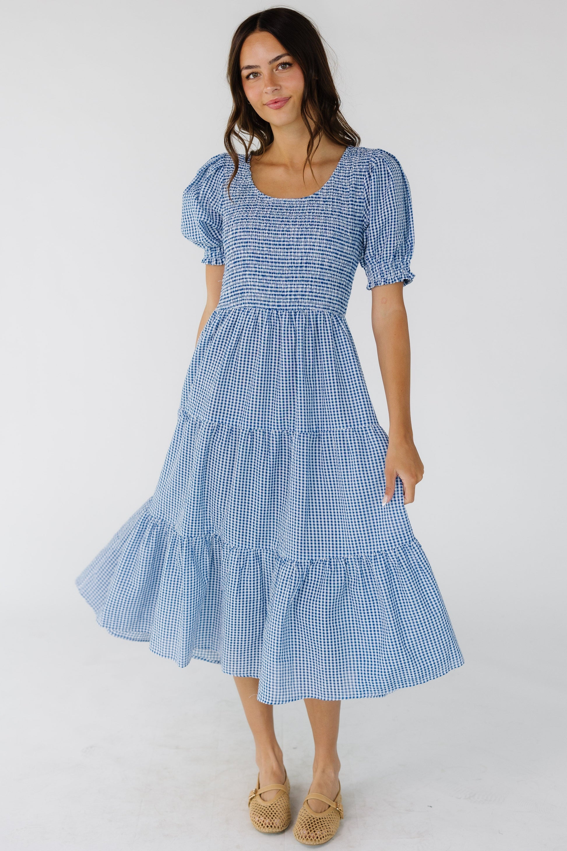 Woman wearing a blue & white gingham midi dress with multiple gathered tiers, smocked front & back bodice, scoop neckline and short puff sleeves.