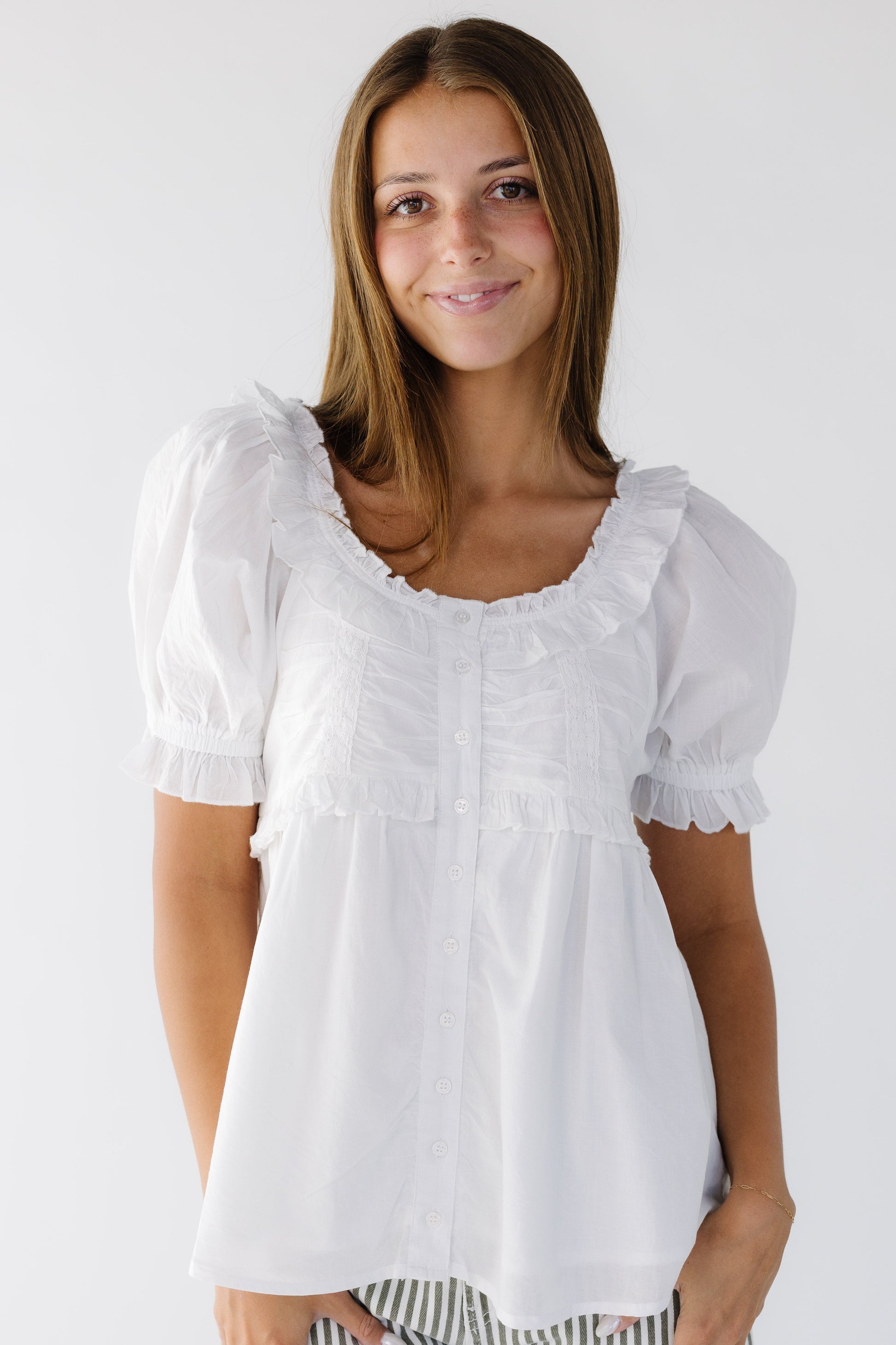 Woman wearing a button down white blouse with a ruffled scoop neckline and short puff sleeves.