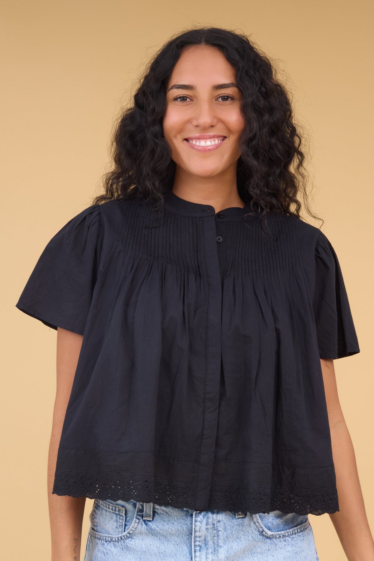 Young woman wearing a black loose fitting top with pintucks, short sleeves and eyelet trim at the hem.