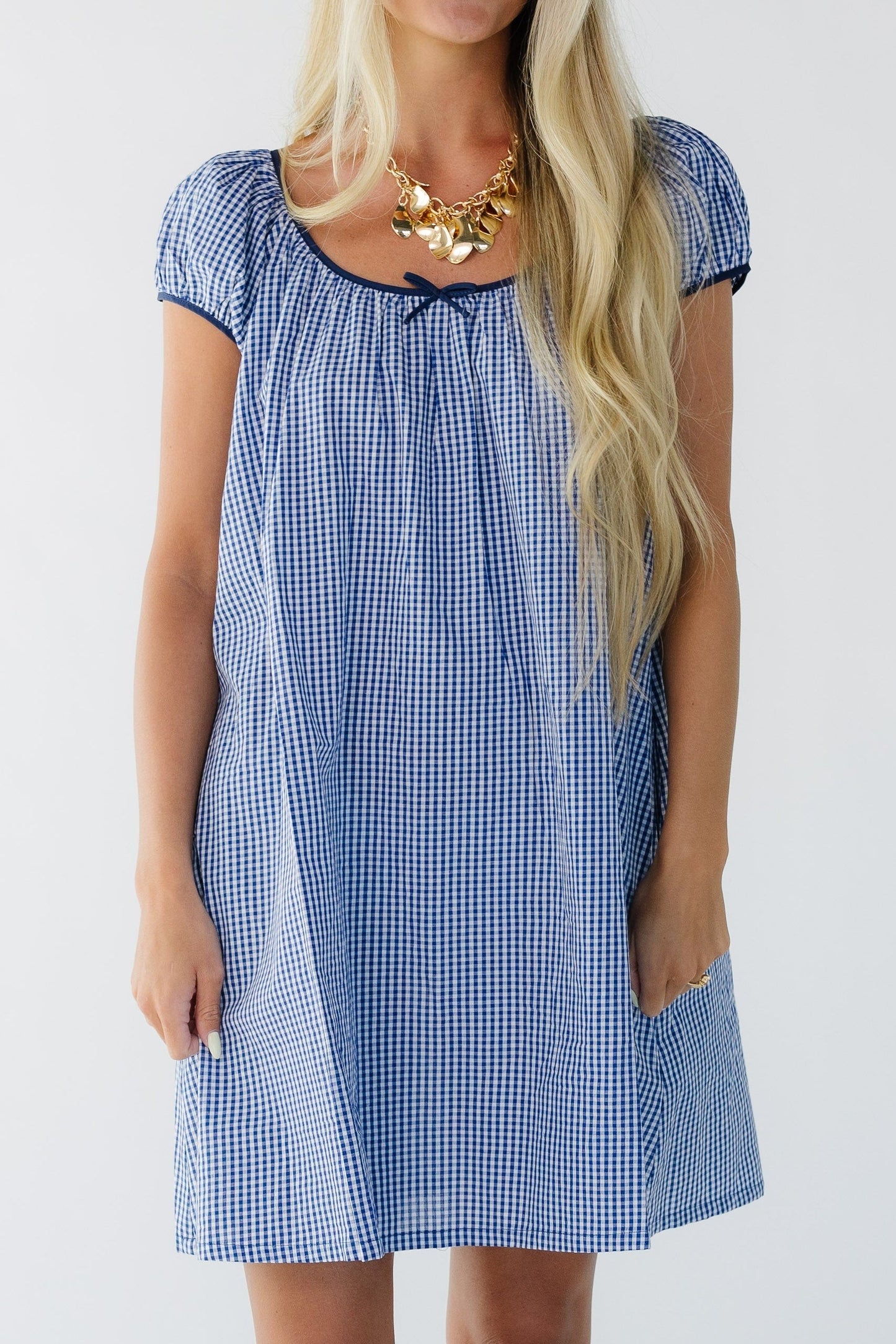 Full dress view of a young woman wearing a short  navy gingham dress with short puff sleeves and a wide scoop neckline.