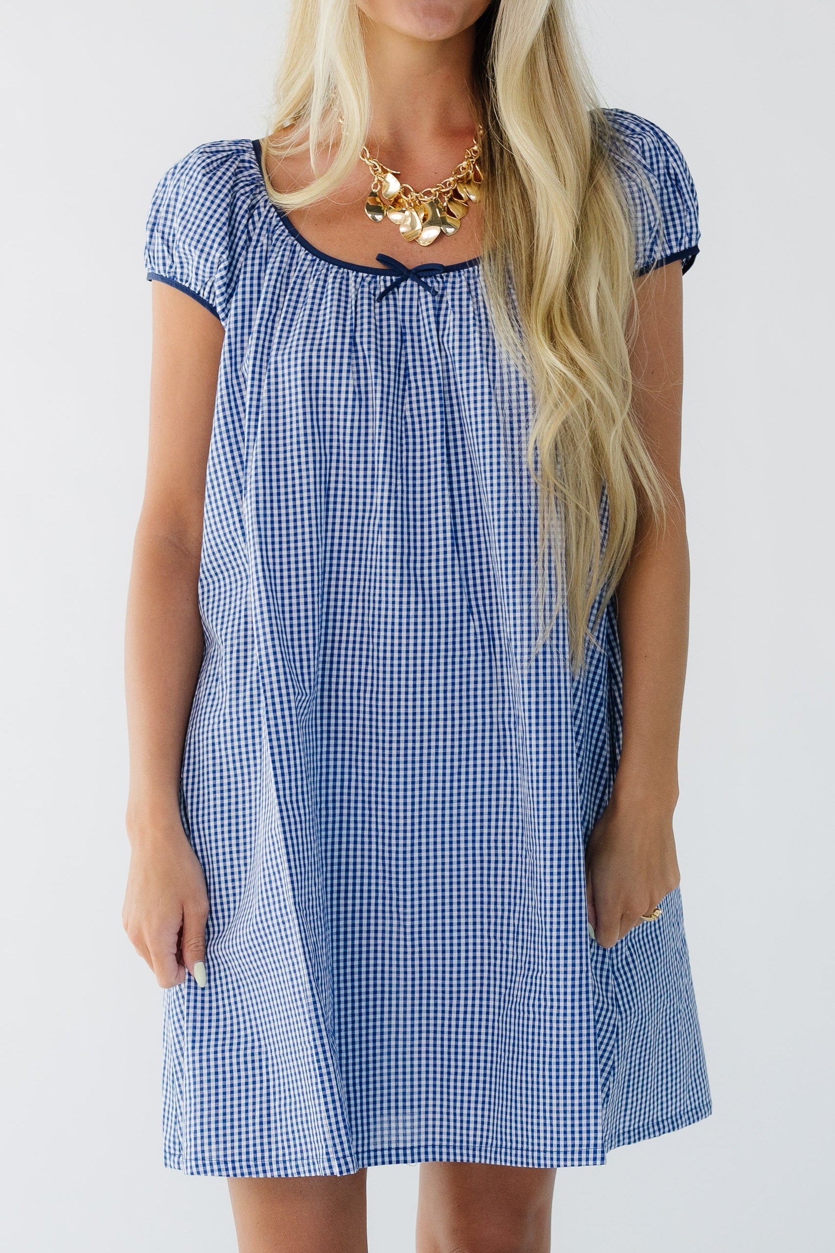 Full dress view of a young woman wearing a short  navy gingham dress with short puff sleeves and a wide scoop neckline.