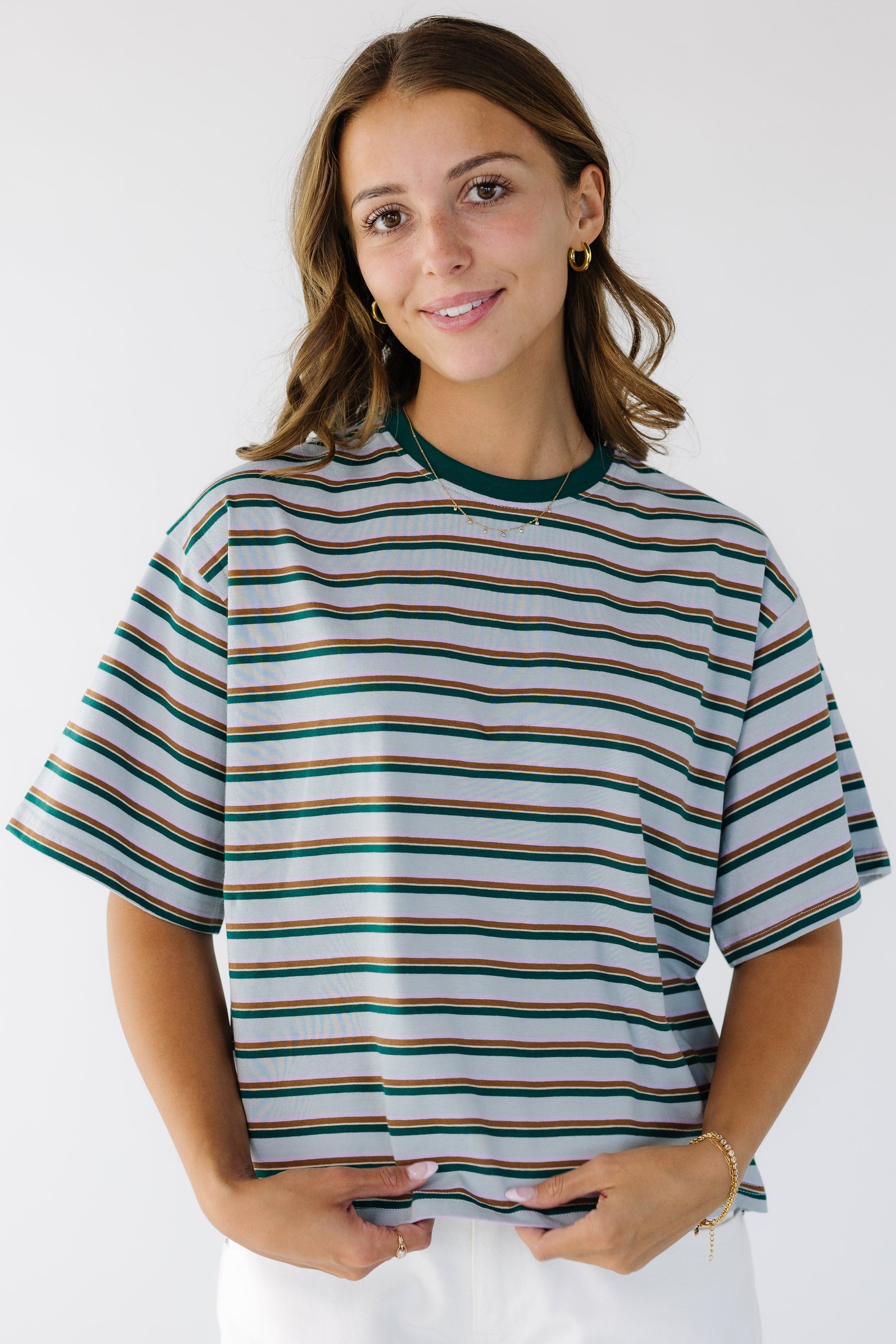 Young woman wearing a  loose fitting stripe tee in blight blue with green & brown stripes and short sleeves.