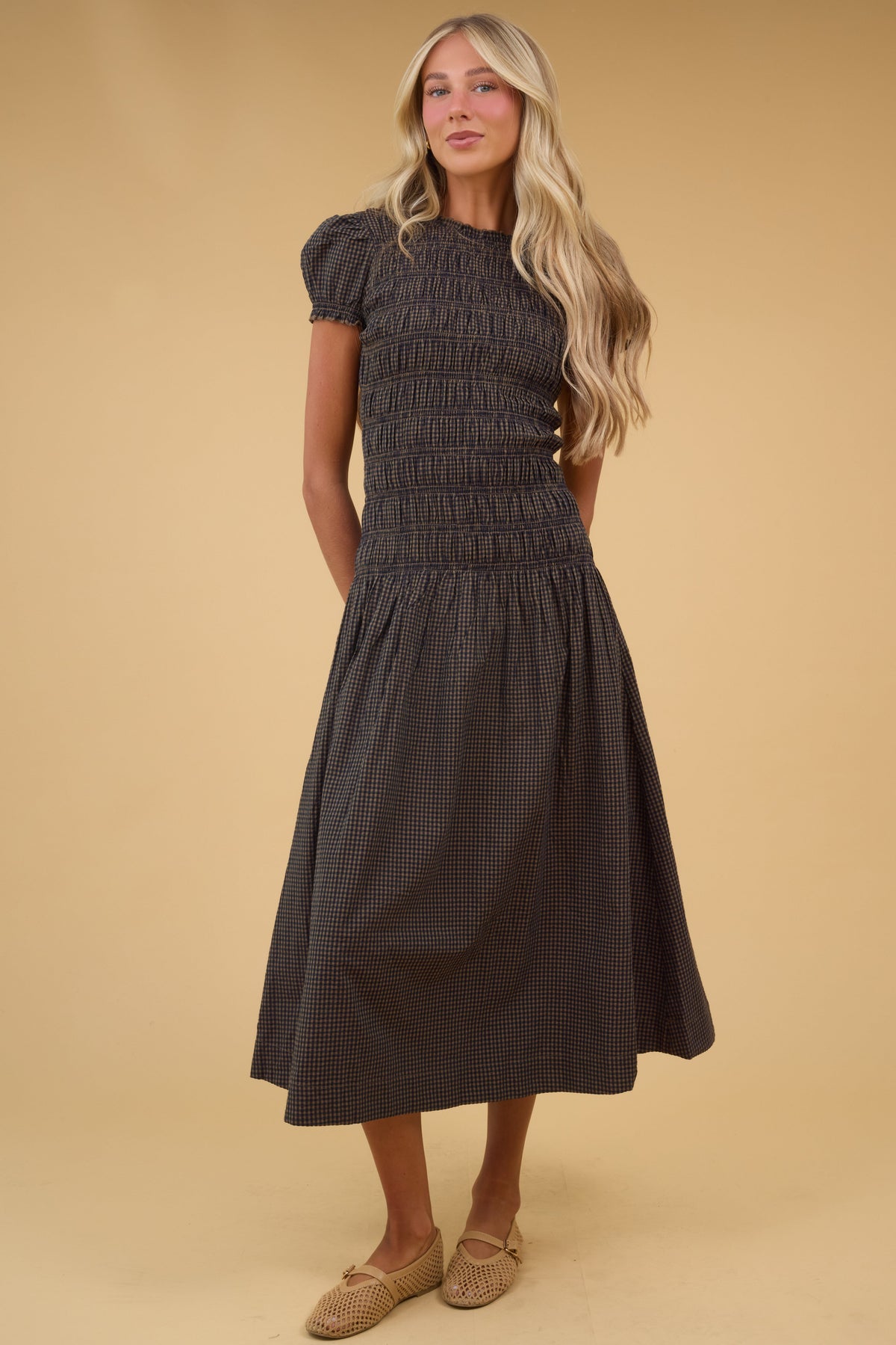 Young woman wearing a modest brown & black gingham midi dress wtih short puff sleeves, round neckline and a smocked bodice the drop waist and a full skirt.