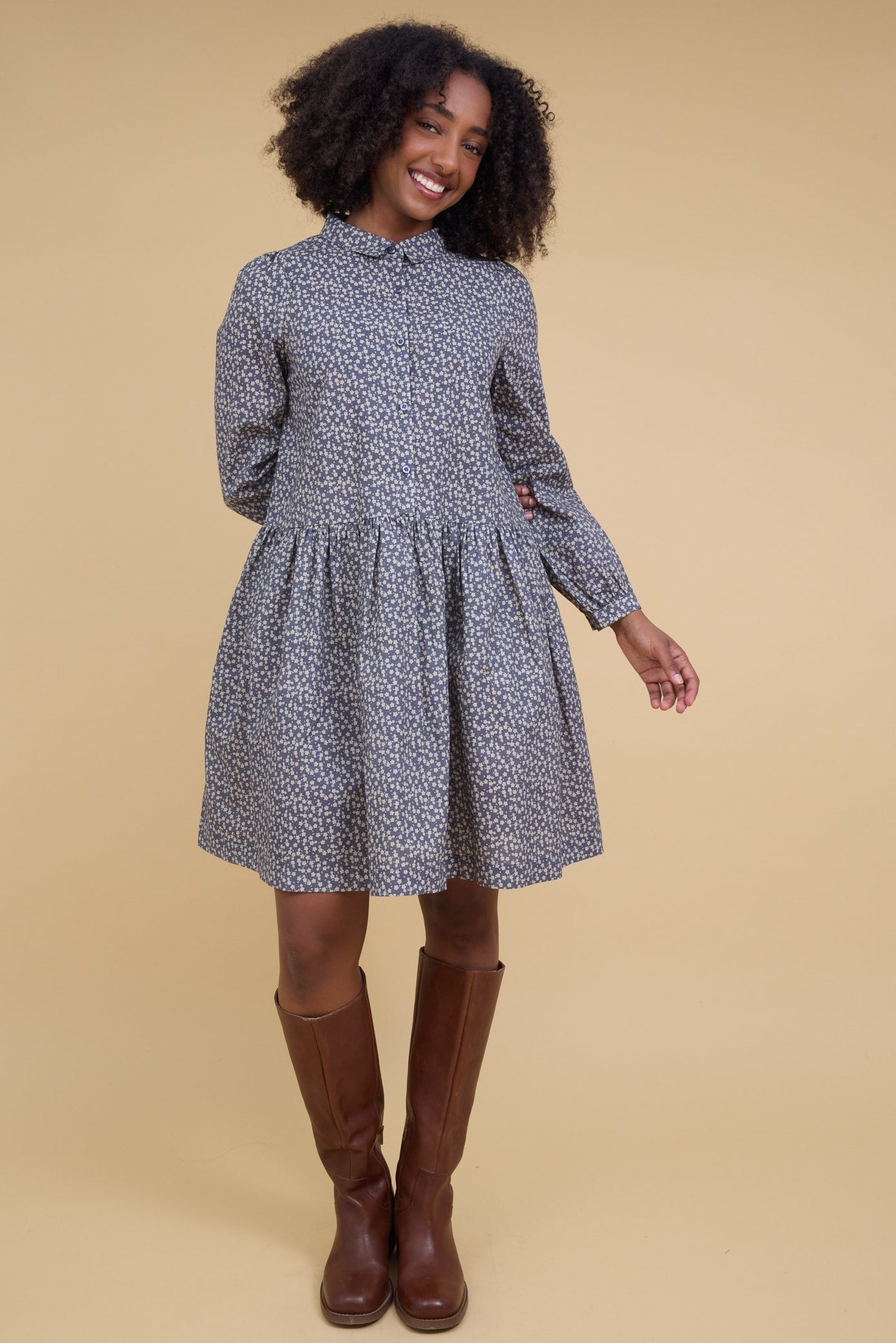 Young woman wearing a charcoal floral print short dress with a rounded neckline & pointed collar, long sleeves and gathered skirt.