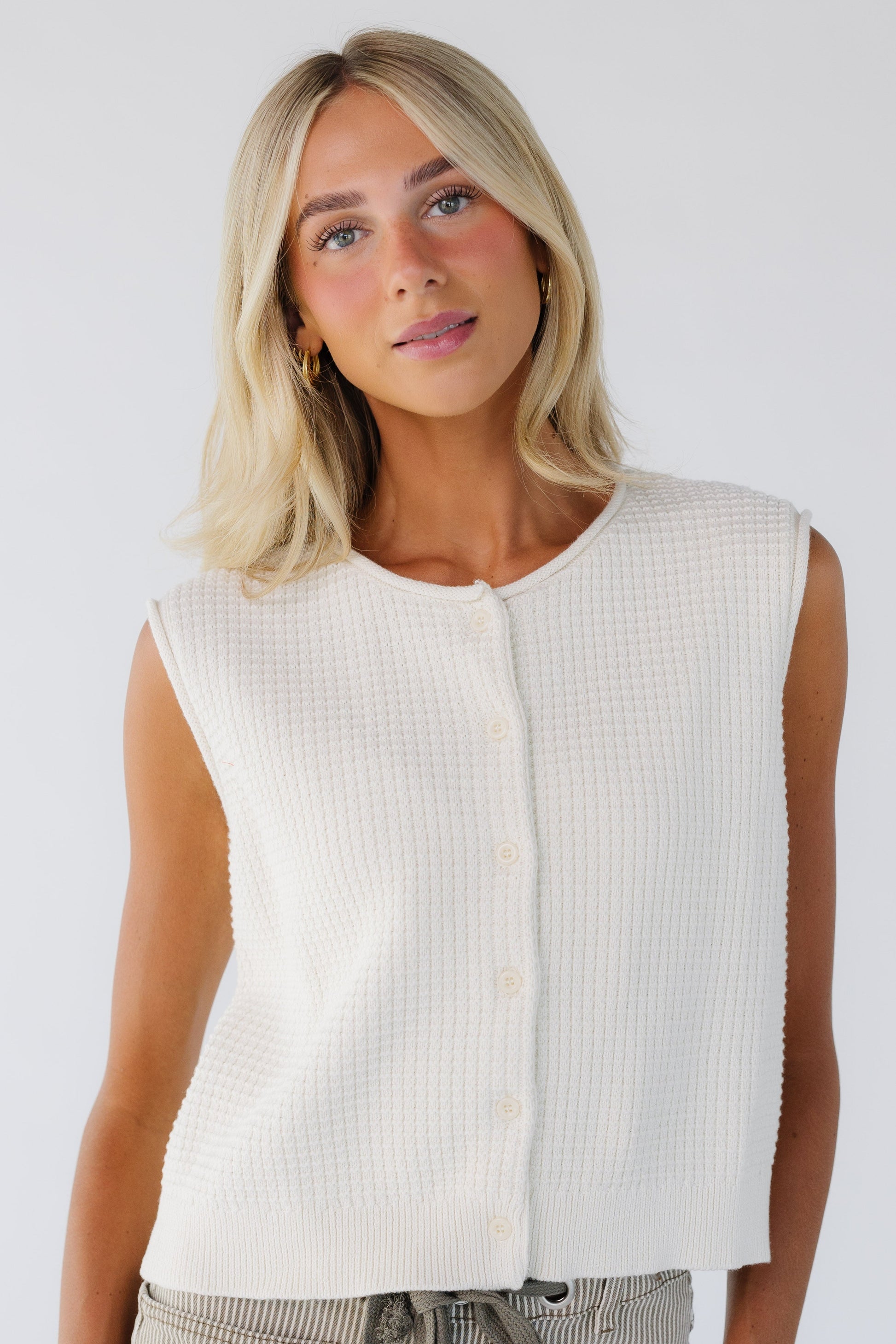 Young woman wearing a cream sleeveless button down sweater vest with a round neckline.