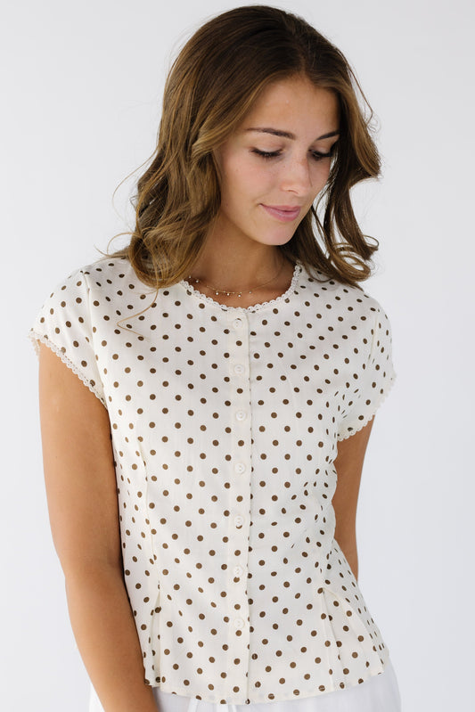 Woman wearing a modest button down blouse in cream and brown polka dots with lace trimmed neckline and sleeves with pleats at the front & back waist.
